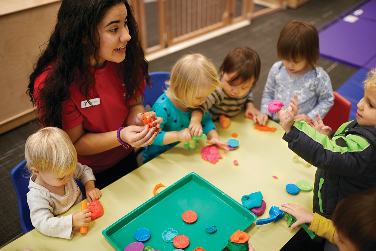 A teacher in a red shirt interacts with a group of young children playing with colorful modeling clay at a table in a classroom.