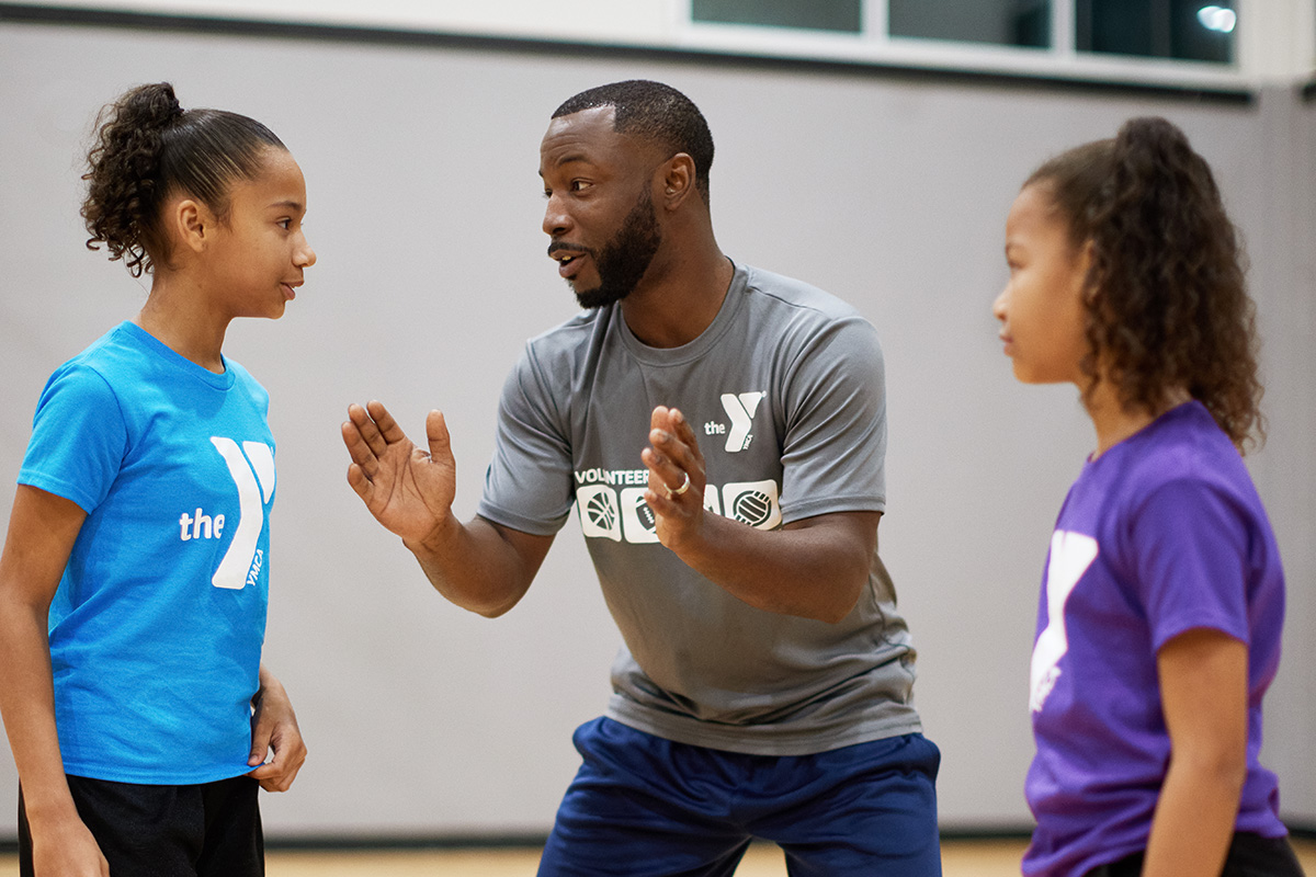 YMCA coach instructing two young girls during a sports practice, engaging them with focus and encouragement in a gymnasium setting.