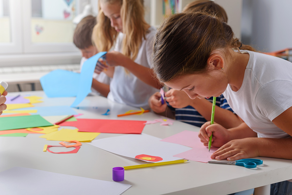 Children work on colorful arts and crafts projects at a table, cutting and drawing with paper and supplies.