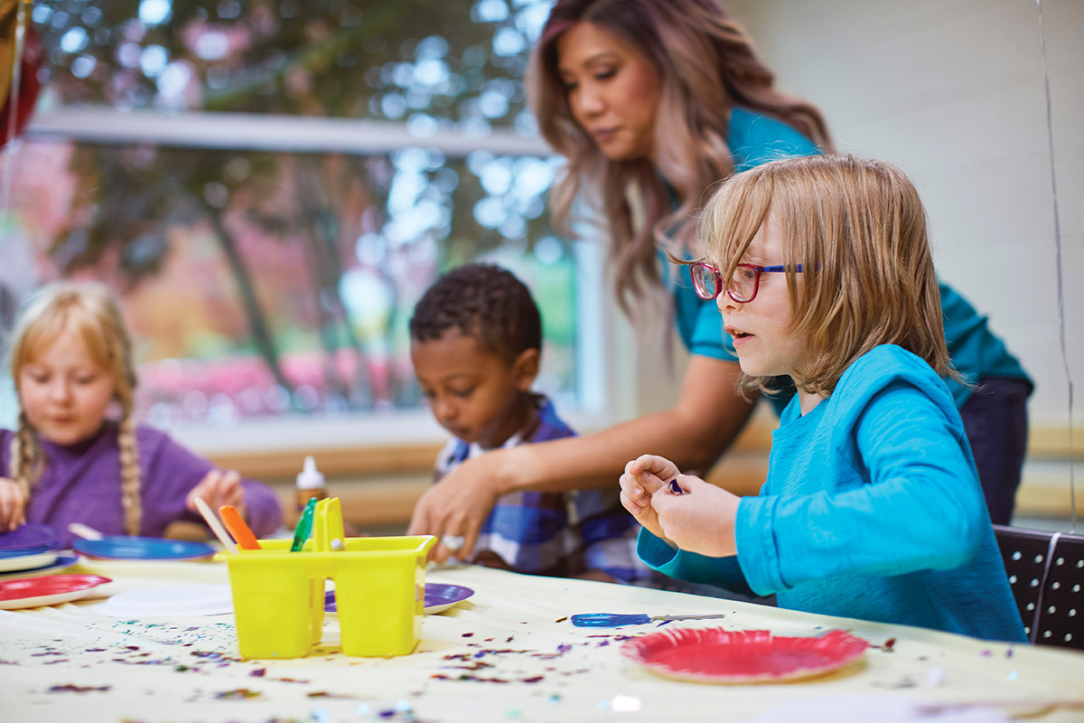 Children engaged in arts and crafts at a table with a teacher assisting in a classroom setting.