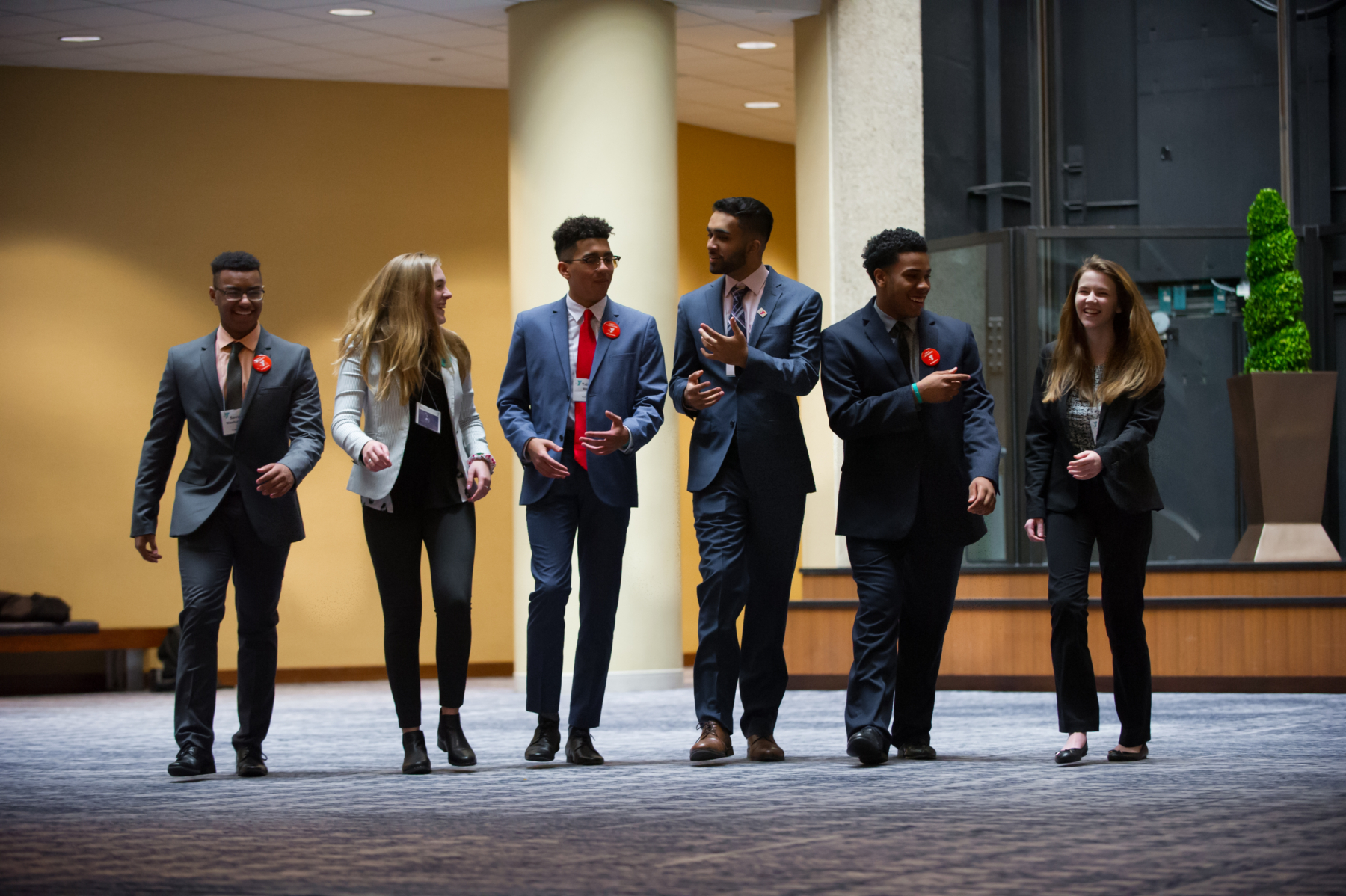 Group of young professionals in formal attire walking together in a conference setting, smiling and engaged in conversation.