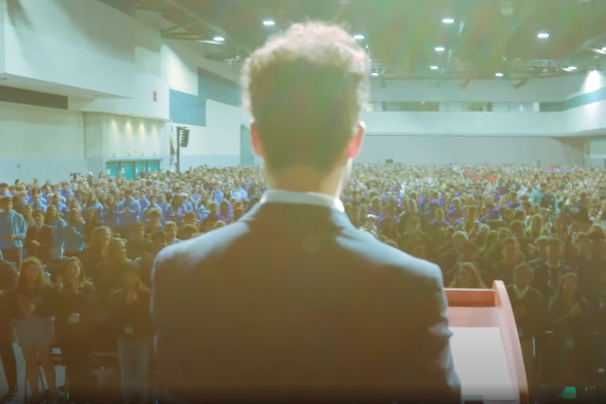 View from behind a speaker at a podium addressing a large audience in a conference hall, with bright lights overhead.