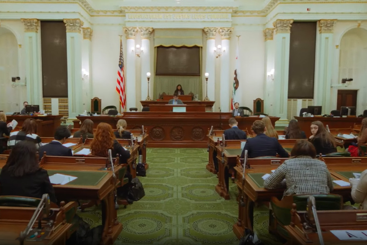 Large legislative chamber with people seated at desks, a speaker at the podium, and flags displayed in a formal setting.