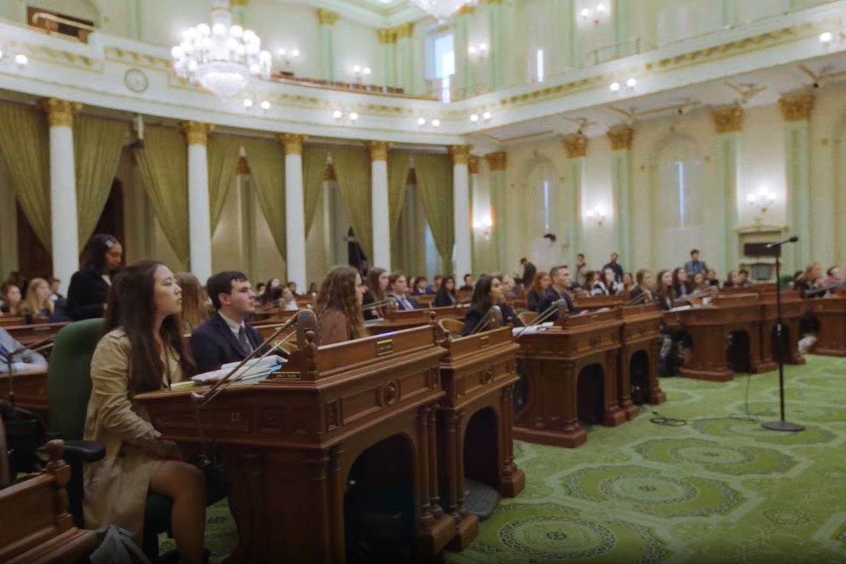 Participants seated at desks in a formal legislative chamber during a youth government event.