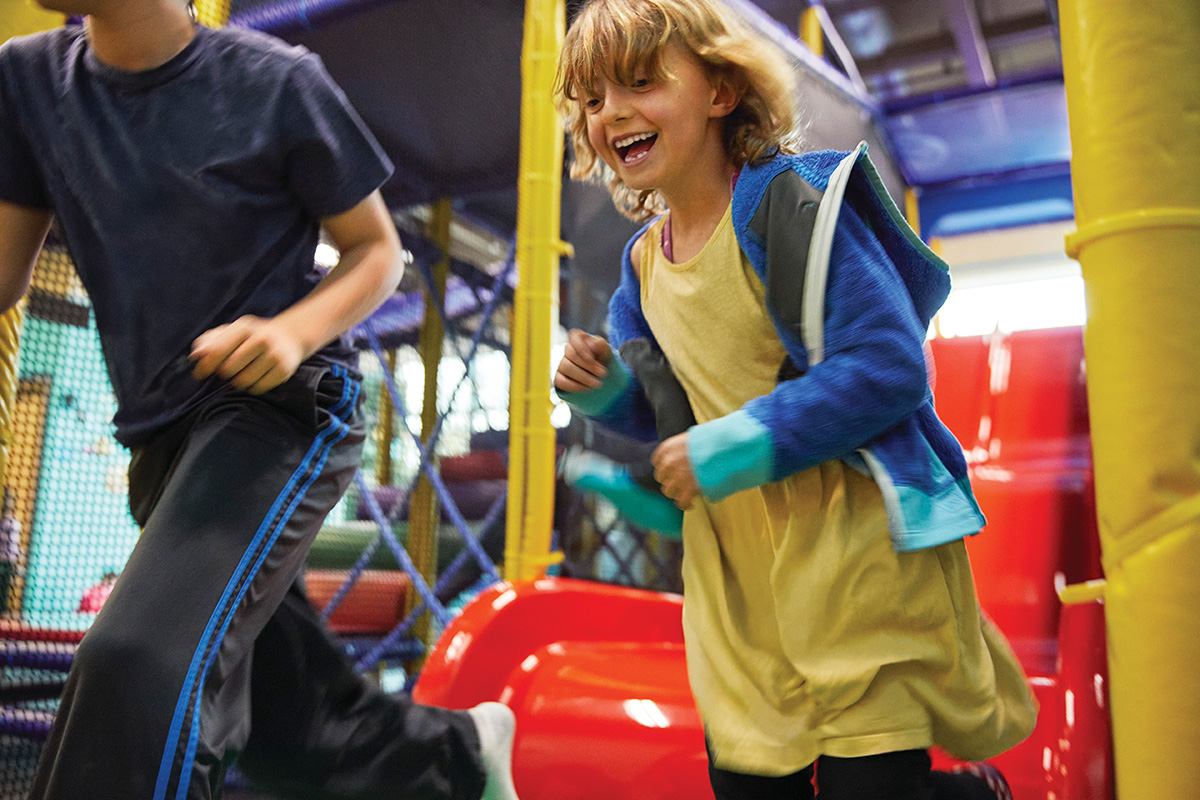 Two children laughing and running in an indoor play area with colorful climbing structures and slides in the background.