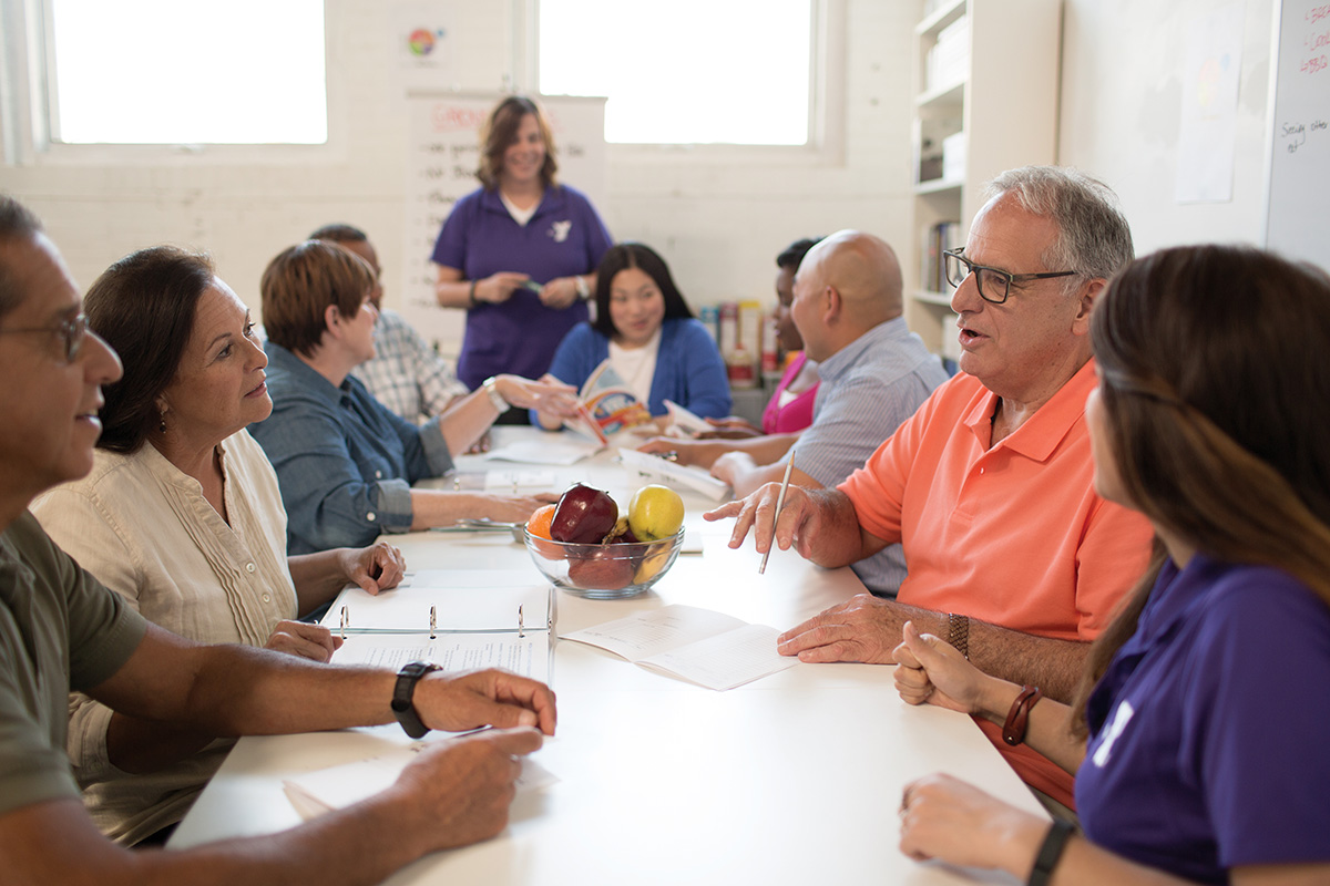 Group of adults participating in a nutrition and wellness class, sitting at a table with materials and a bowl of fruit.