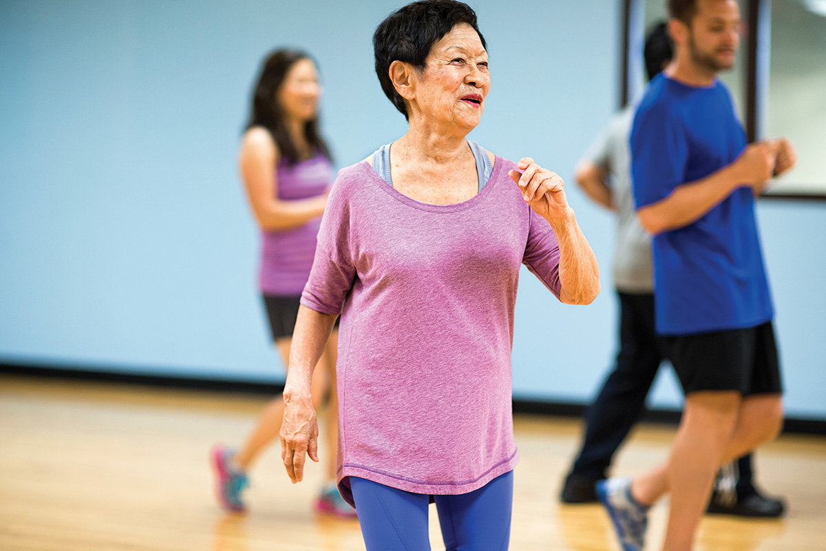 An older woman in a purple top participating in a group fitness class, smiling and moving in a bright indoor studio.