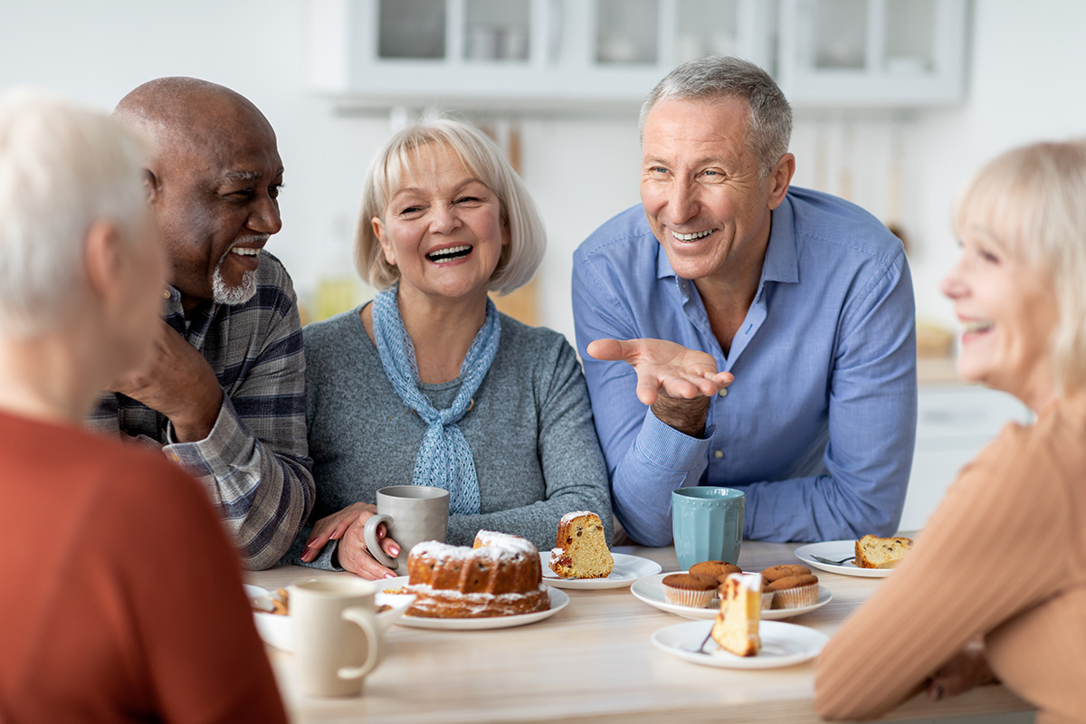 Group of older adults laughing and enjoying coffee and desserts together at a table in a bright kitchen setting.