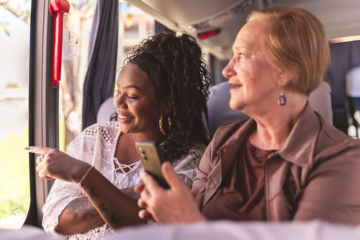 Two women sitting on a bus, one pointing out the window while the other smiles and holds a smartphone, engaged in conversation during a trip.