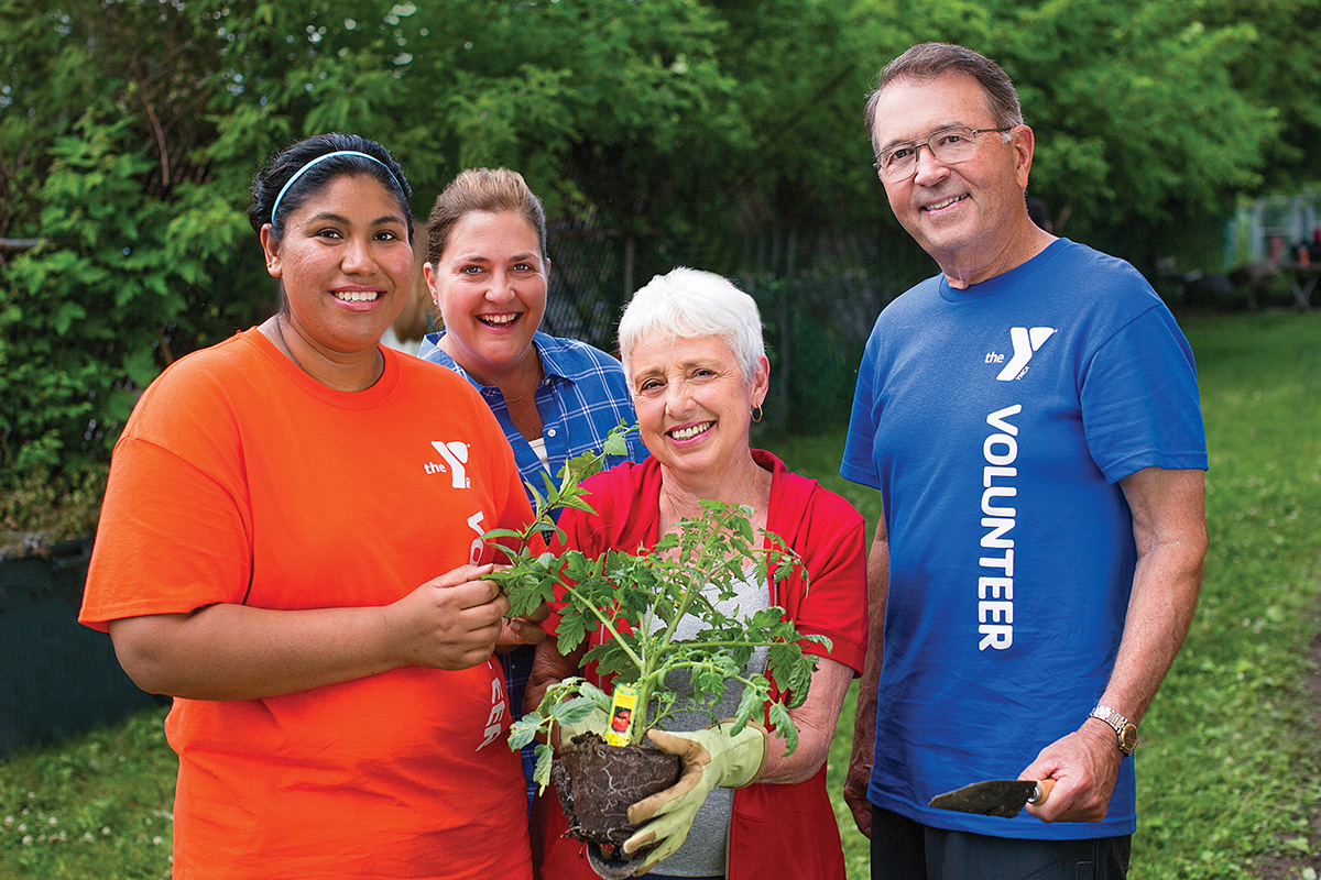 Group of volunteers smiling and holding gardening tools and plants in an outdoor setting, wearing YMCA-branded shirts.