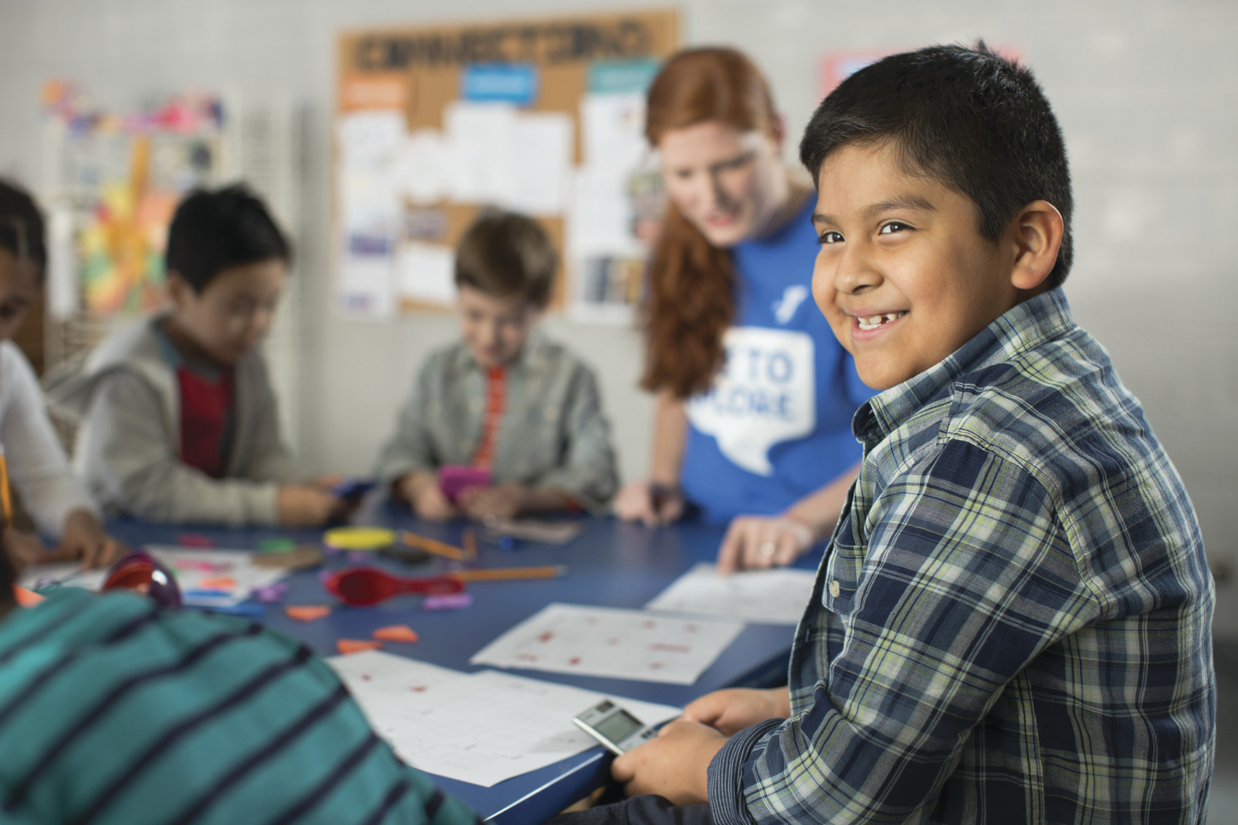 Smiling boy in a classroom setting with other children and a teacher working on crafts at a table.