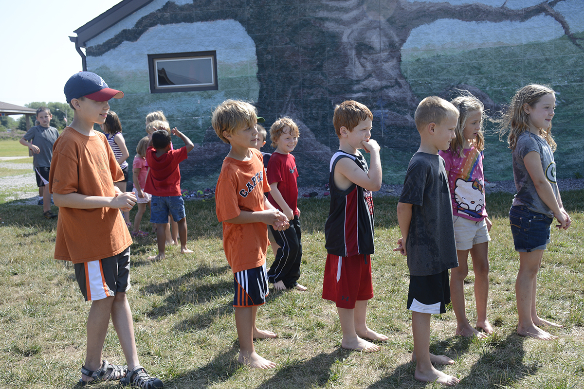 Children standing in a line outdoors on a sunny day, participating in an activity near a mural of a tree painted on a building.