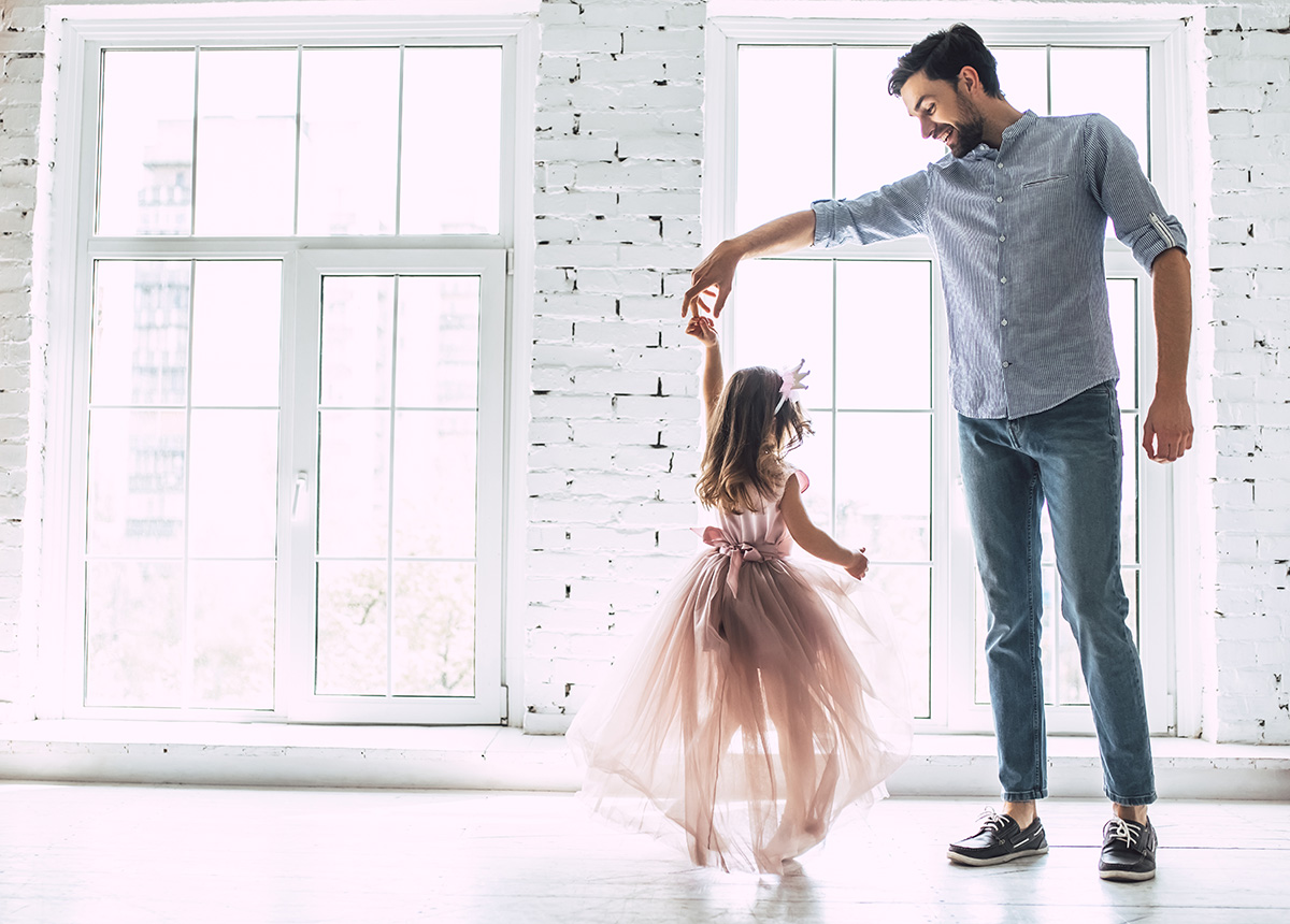 A father twirls his young daughter dressed in a flowing pink dress in a bright room with large windows and white brick walls, creating a joyful and tender moment.