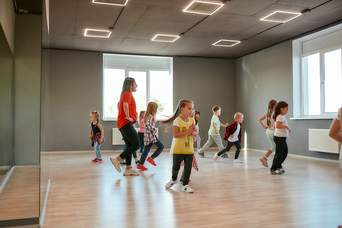 Group of children in a dance studio practicing choreography with an instructor, bright room with modern lighting.