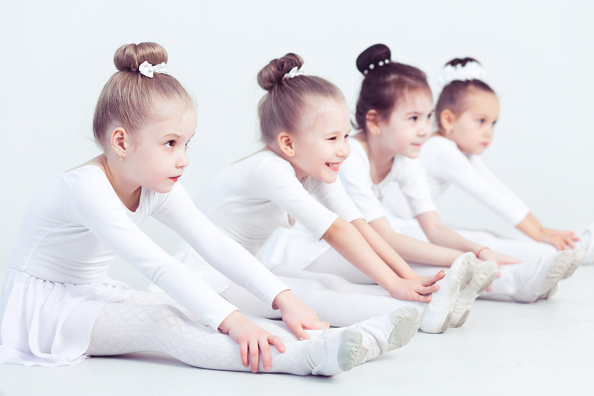 Four young girls in white ballet outfits sitting on the floor, stretching with smiles during a dance class.