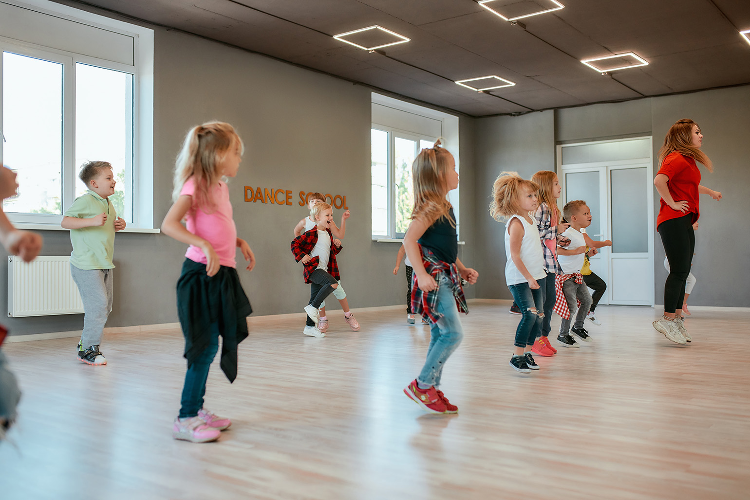 Children participating in a dance class, following an instructor in a brightly lit studio with 'Dance School' signage on the wall.