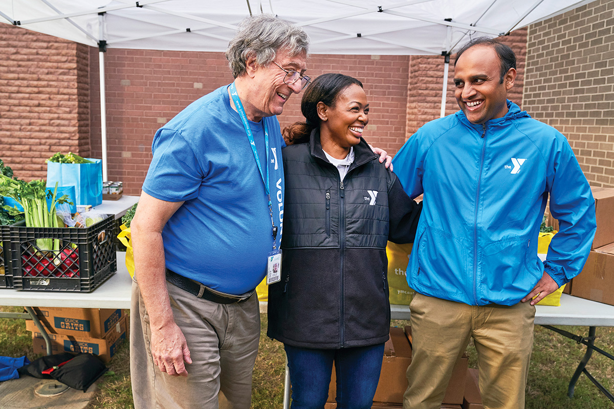 Three YMCA volunteers smiling and standing together at an outdoor food distribution table under a canopy.