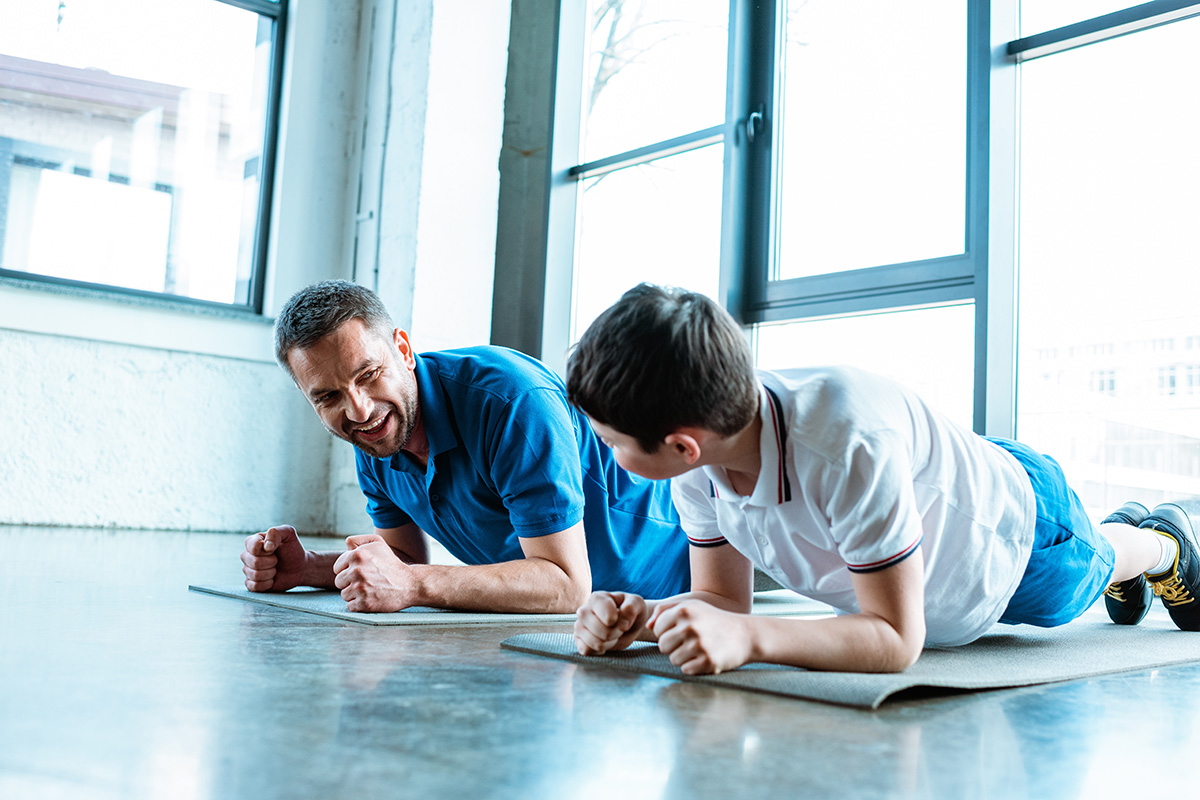 A man and a boy lie in a plank position on yoga mats, smiling and interacting during a workout session in a bright, sunlit room with large windows.