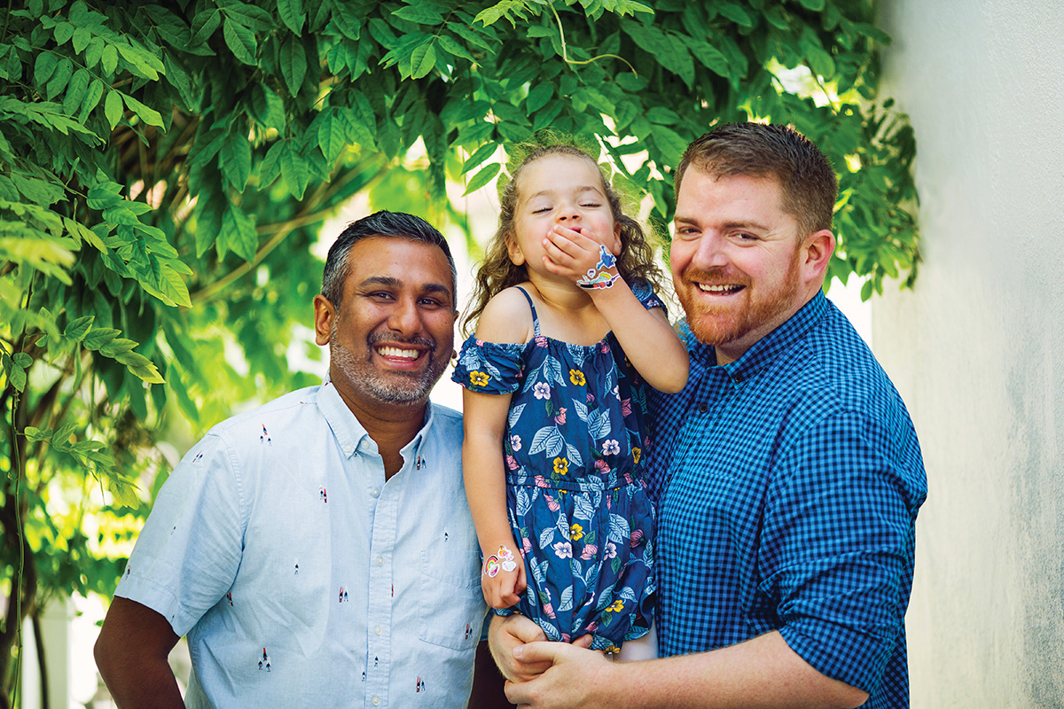 A joyful family portrait featuring two fathers and their young daughter outdoors. The child is laughing, wearing a floral dress, and is held by one father, while both men smile warmly under lush green foliage. Bright natural light enhances the vibrant setting.