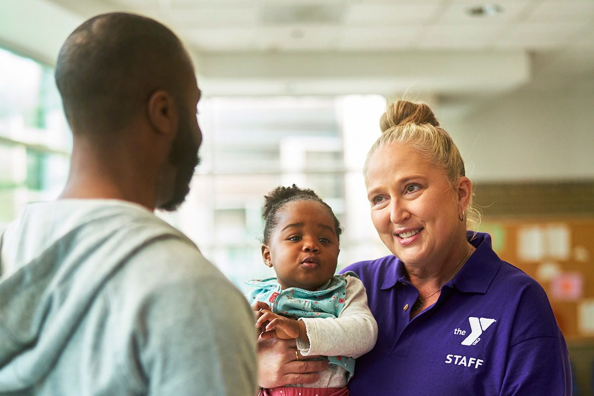 YMCA staff member in purple shirt smiling while speaking with a parent holding a baby.