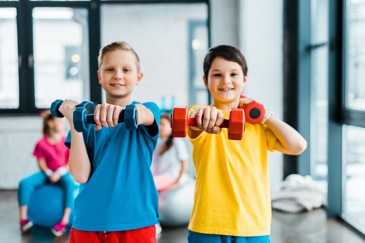 Two boys smiling and holding dumbbells in a fitness class, with children exercising in the background in a bright indoor space.