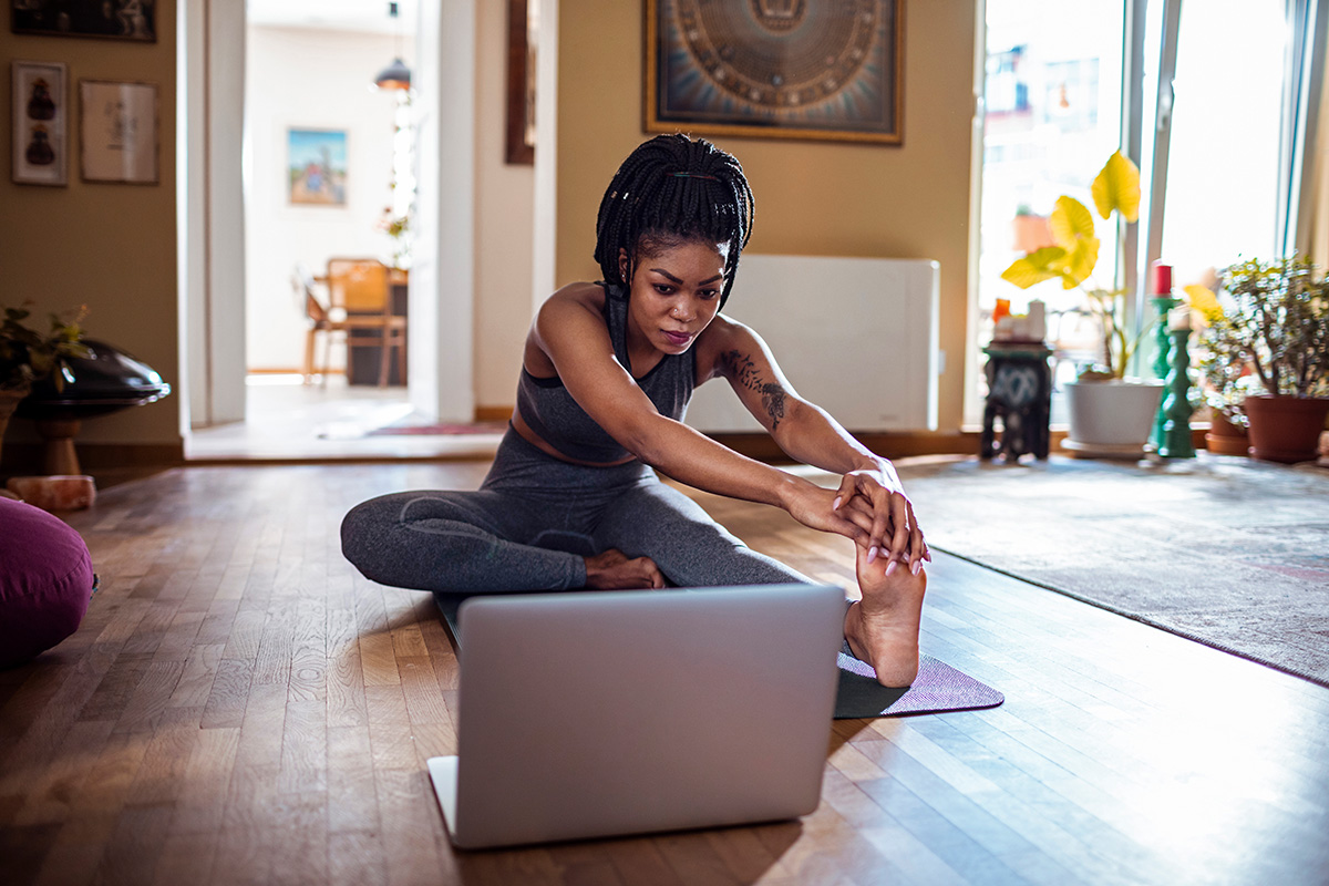 A woman practices yoga at home, stretching on a mat while following a virtual class on a laptop in a brightly lit room.