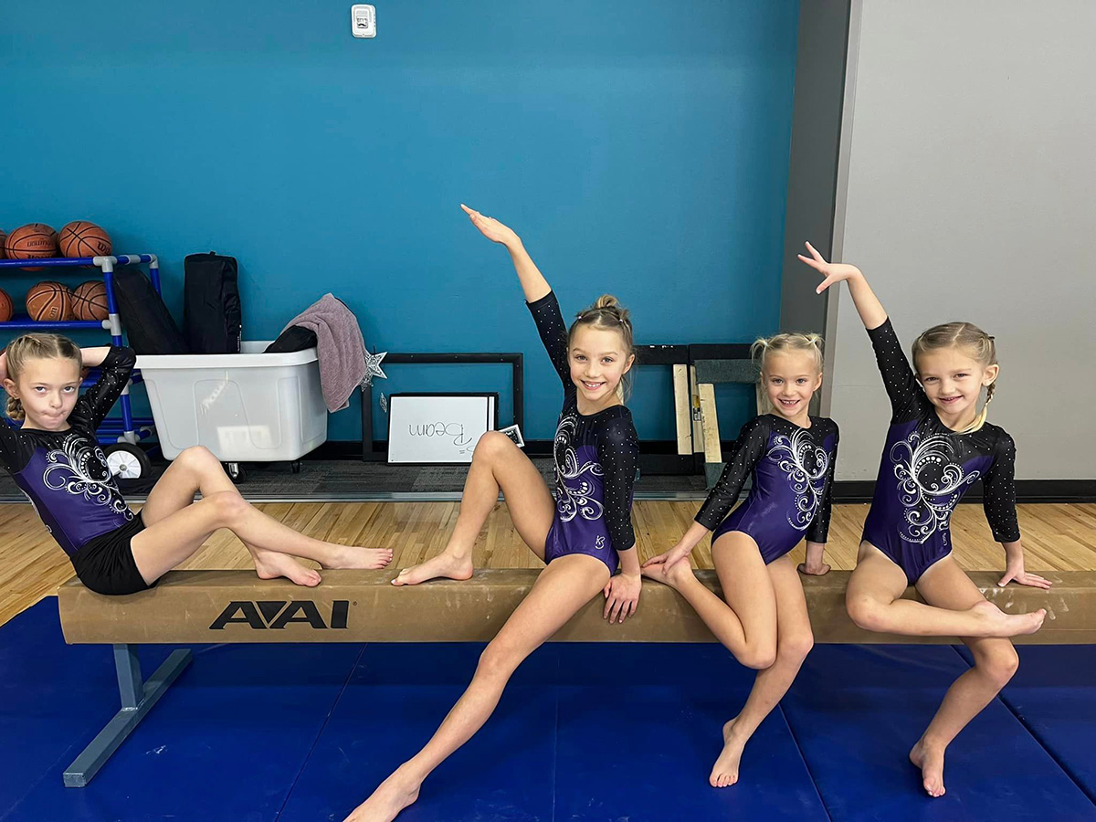 Four young gymnasts in purple and black leotards posing on a balance beam in a gym with a blue wall.