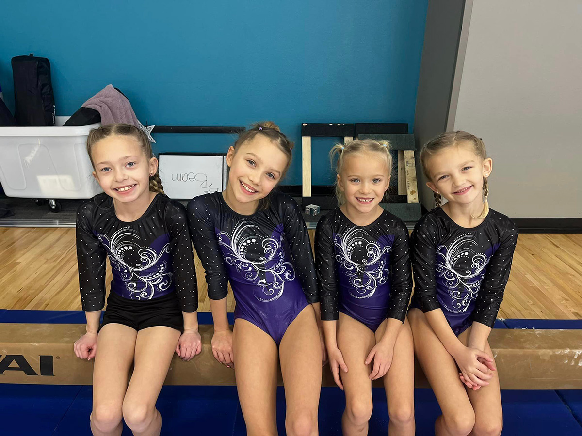 Four young gymnasts in matching leotards sitting on a balance beam, smiling at the camera inside a gym.