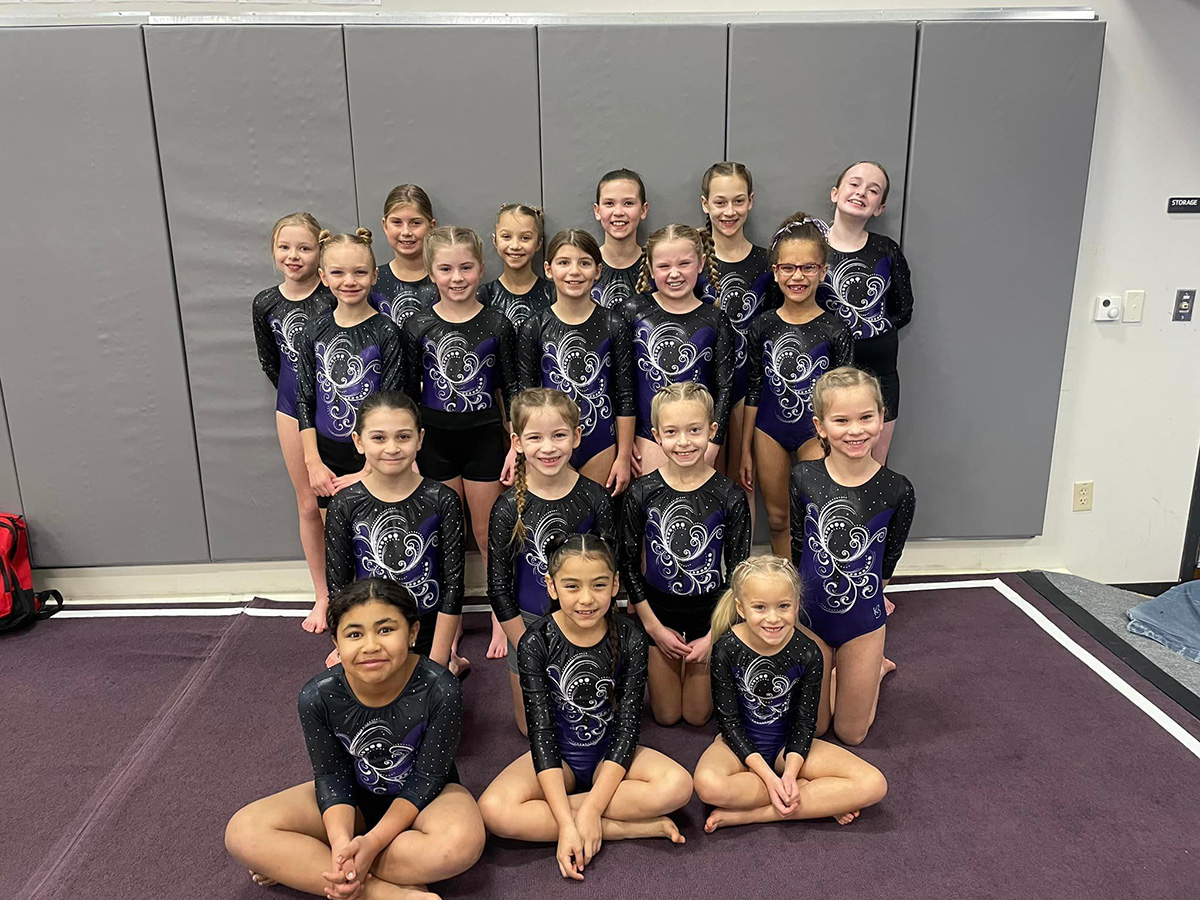 Group of young gymnasts in matching purple and black leotards posing together on a gym floor.