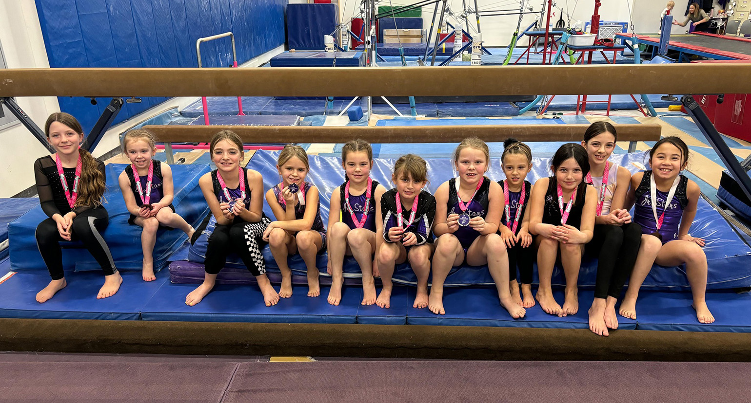 Group of young gymnasts sitting together on mats in a gymnastics facility, wearing medals and smiling at the camera.