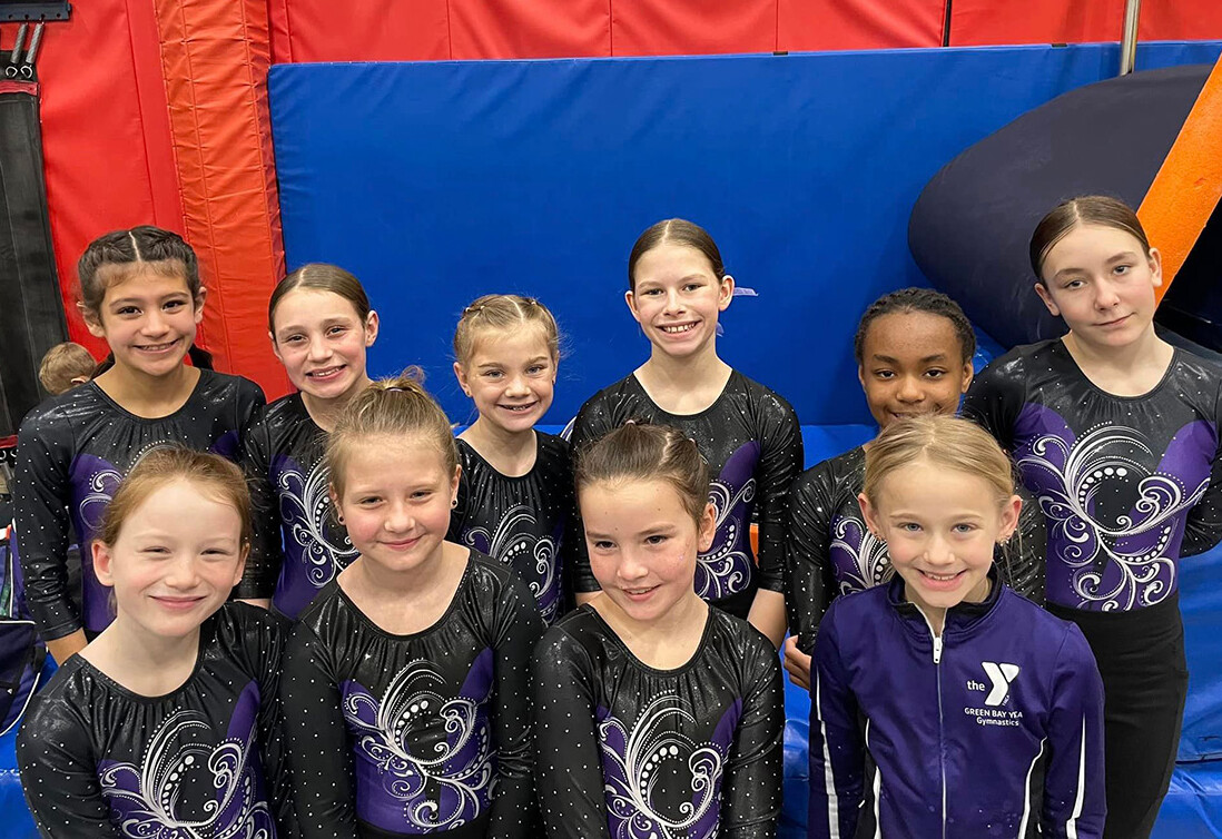 A group of young gymnasts in matching black and purple leotards smile for a photo in a gymnastics gym with colorful mats and equipment in the background.