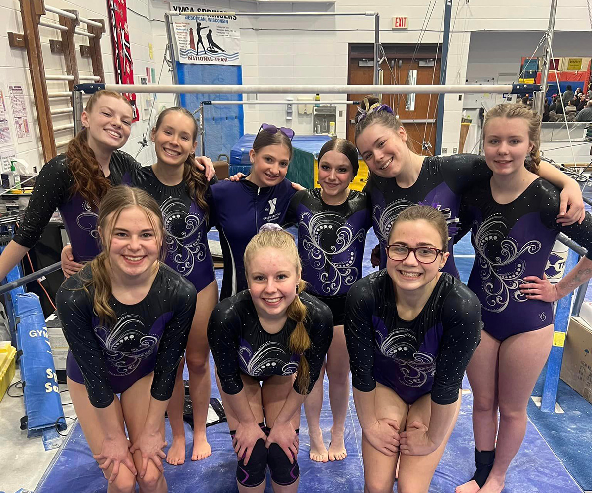 A group of smiling gymnasts wearing matching black and purple leotards posing together in a gymnastics facility with equipment in the background, showcasing their camaraderie and team spirit.