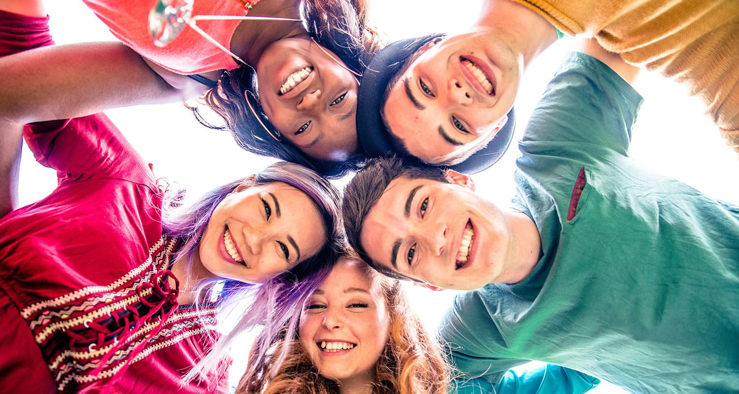 A group of five smiling friends huddle together, looking down at the camera in a bright, cheerful setting.