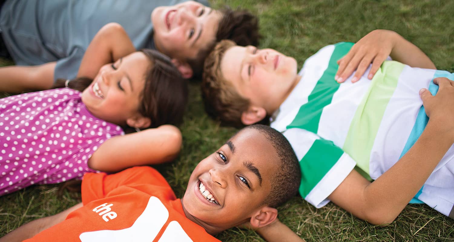 Four smiling children lying on grass in a circle, relaxing outdoors, with one child wearing an orange YMCA shirt in the foreground.