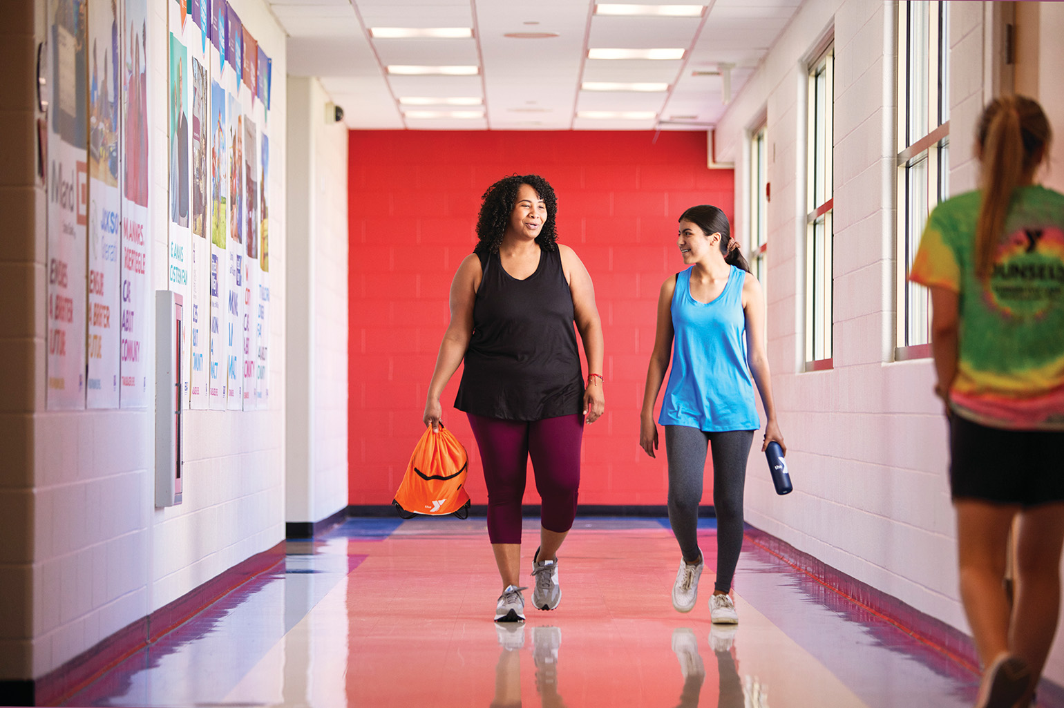 Two women in athletic wear walk down a brightly lit hallway with posters on the walls, carrying a gym bag and water bottle.