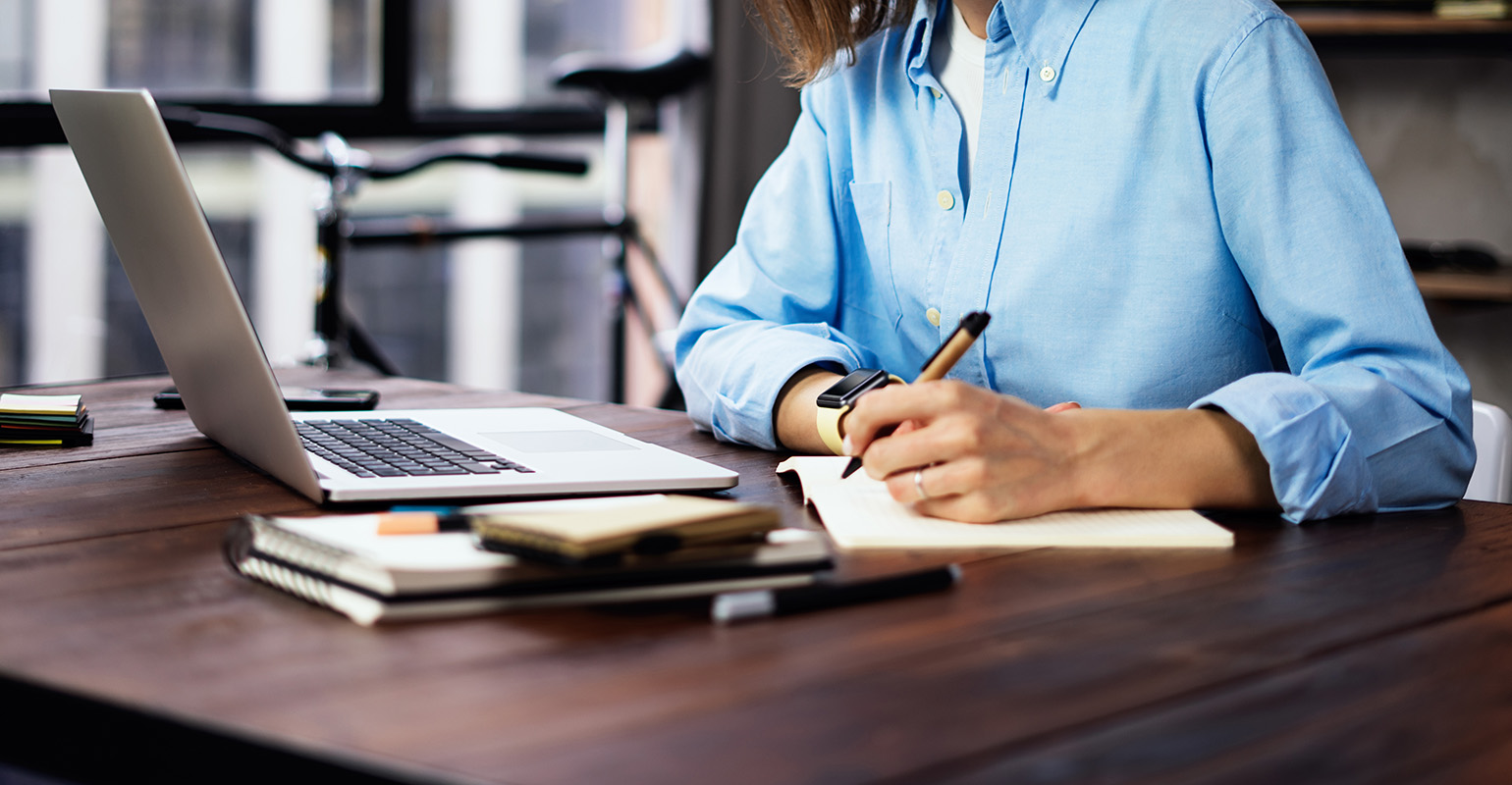 A person in a blue shirt writes in a notebook while working on a laptop at a desk with books and a bicycle in the background.