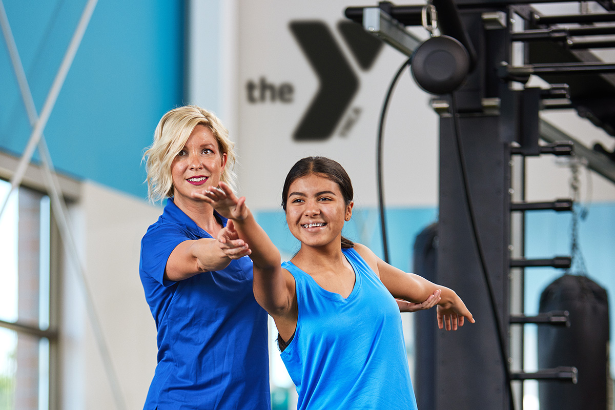 A fitness instructor in a blue shirt guides a smiling young girl during a workout session at a YMCA facility.