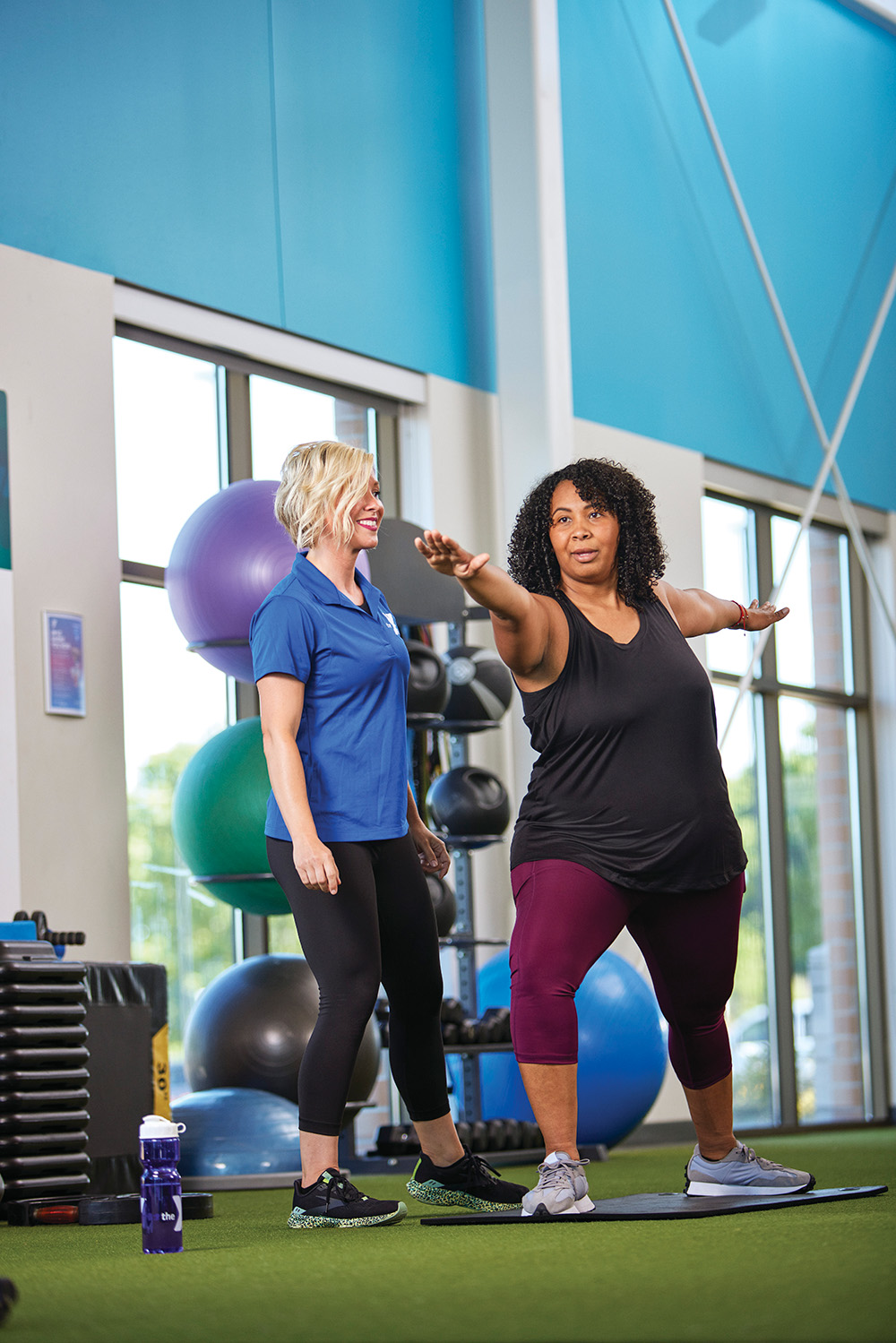 A fitness instructor guides a woman in a Warrior II yoga pose on a turf floor in a gym.