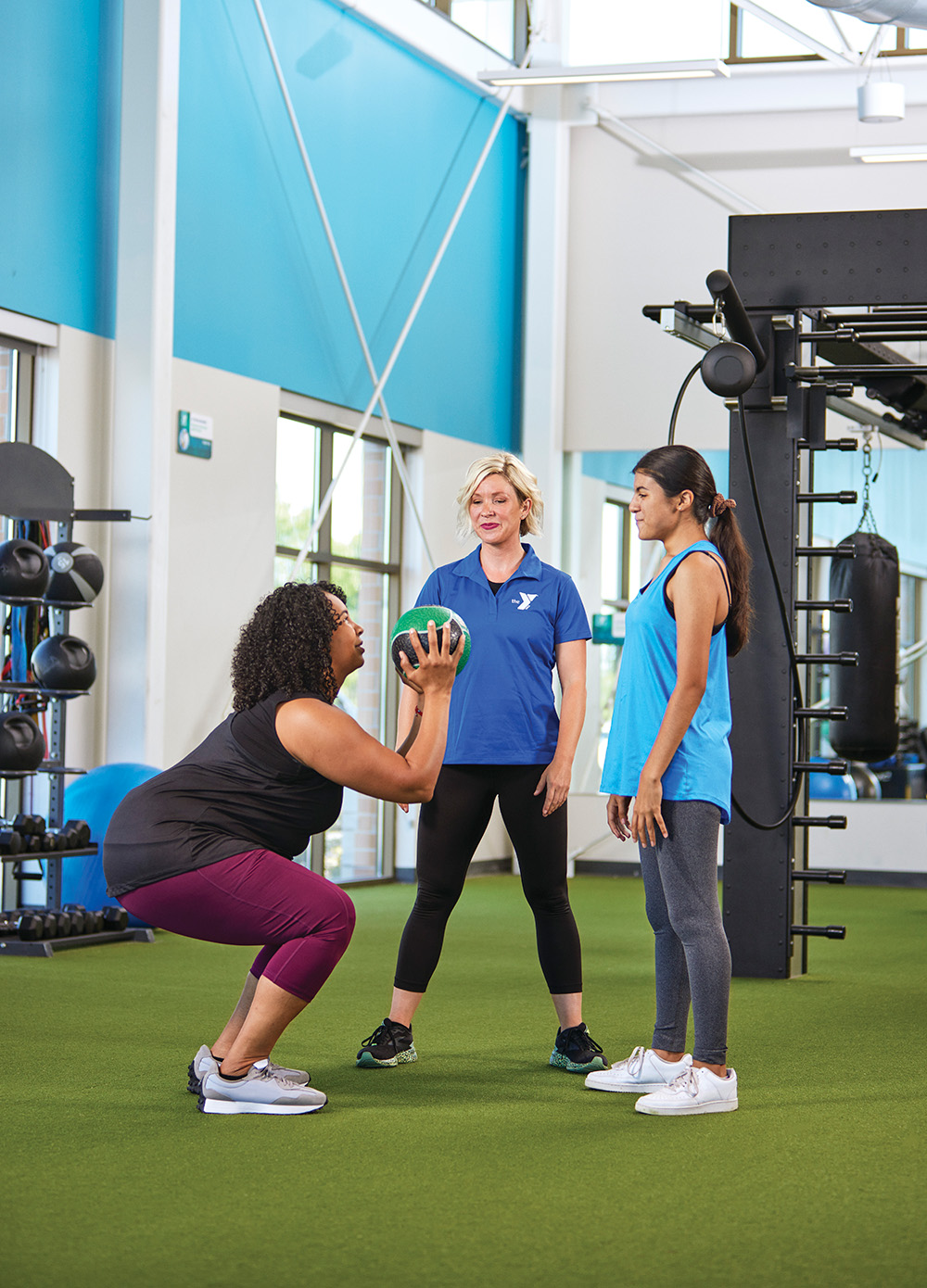 A fitness instructor guides a woman doing squats with a medicine ball as another participant watches in a gym.