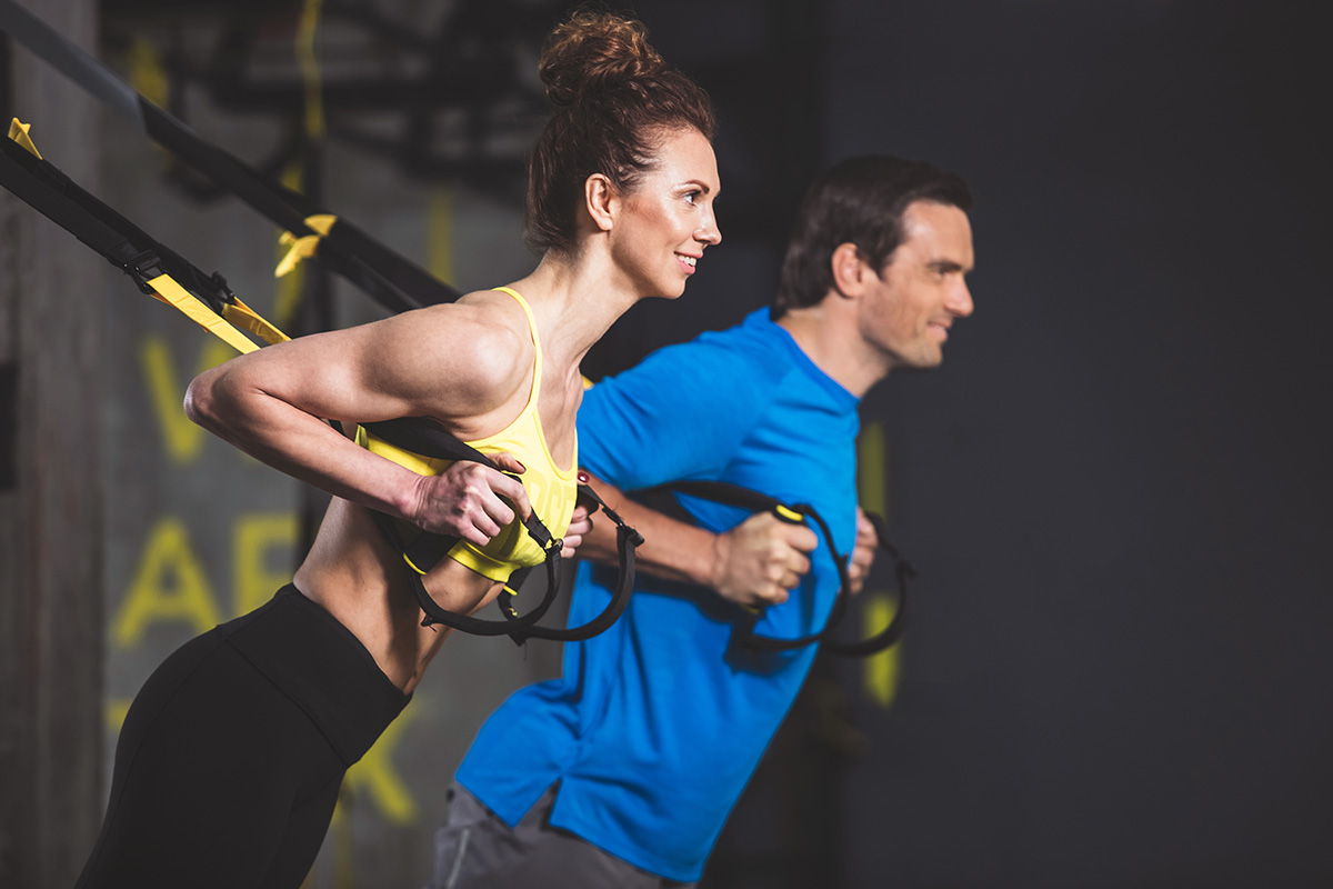 A man and woman perform chest presses using suspension straps during a workout in a gym.