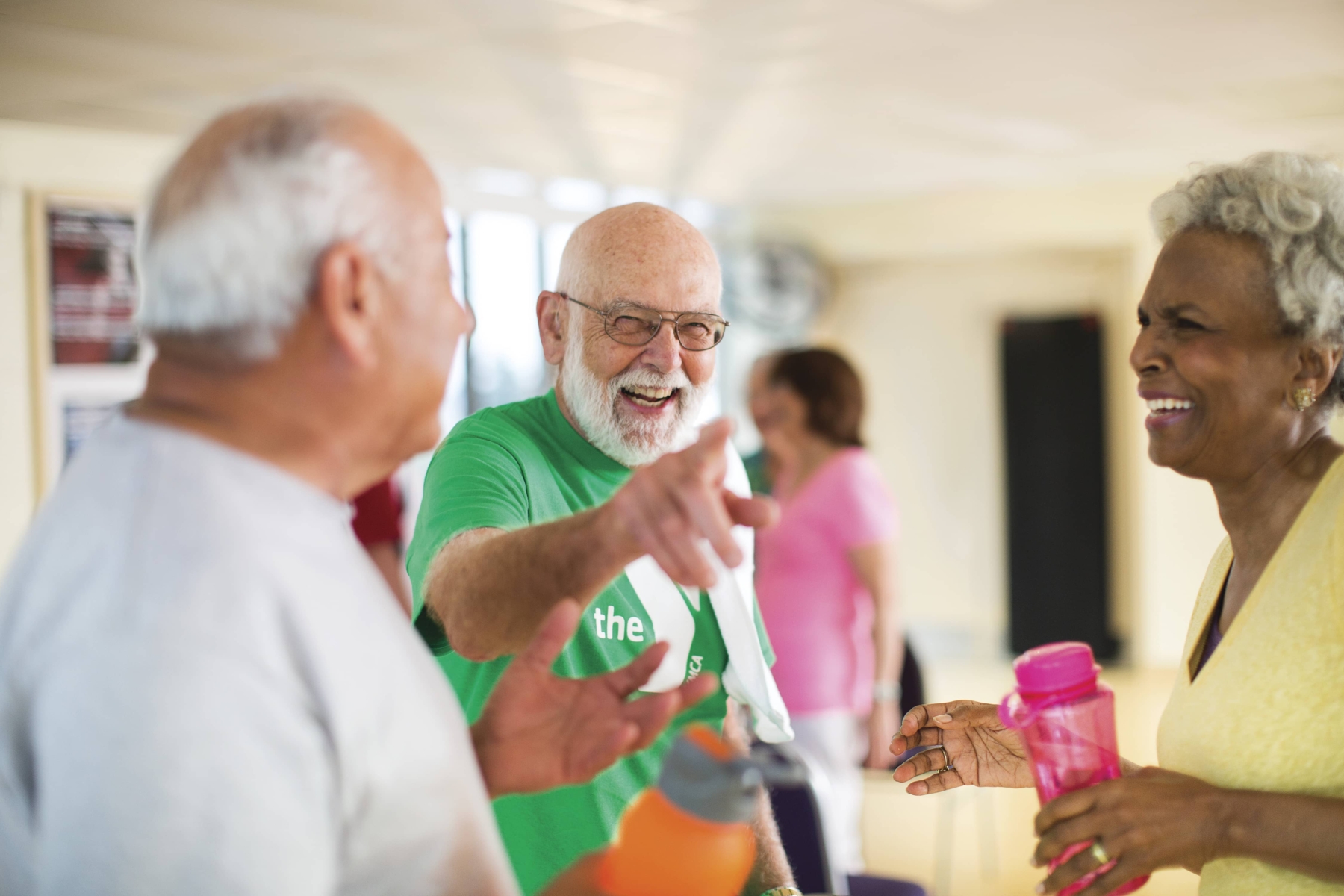 A group of older adults laughing and chatting in a brightly lit room. One man wearing a green shirt points playfully while holding a water bottle. A woman with gray hair holds a pink water bottle, smiling warmly.