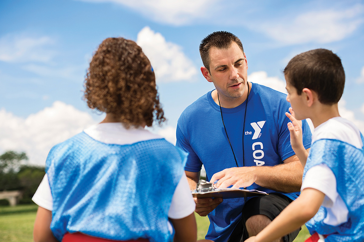 A coach in a blue shirt with a clipboard talking to two young players on a sunny day at a sports field.