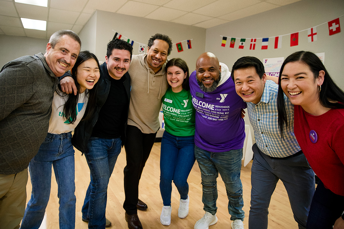 A diverse group of adults smiling and posing together in a community center with international flags in the background.