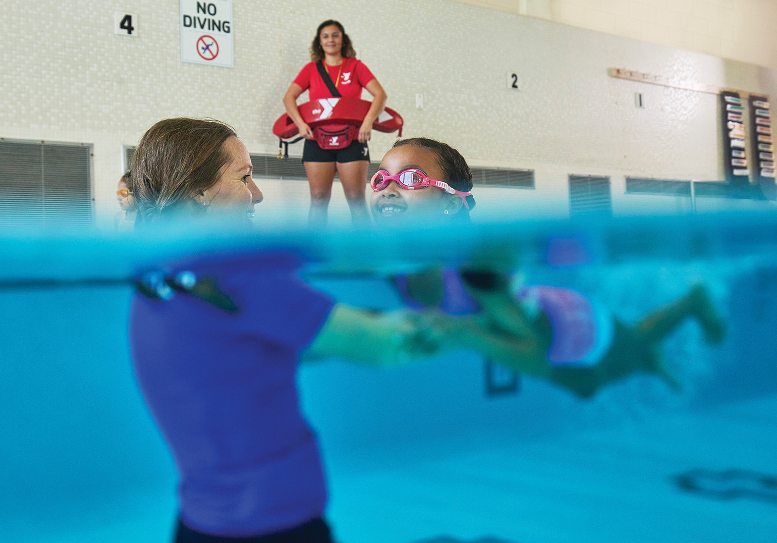 A swim instructor helps a young girl in goggles practice swimming while a lifeguard observes from the pool deck.