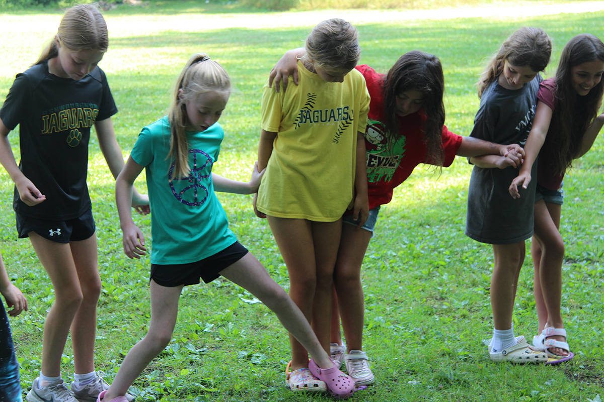 A group of girls works together on a team-building activity outdoors, balancing with linked arms on a grassy field.