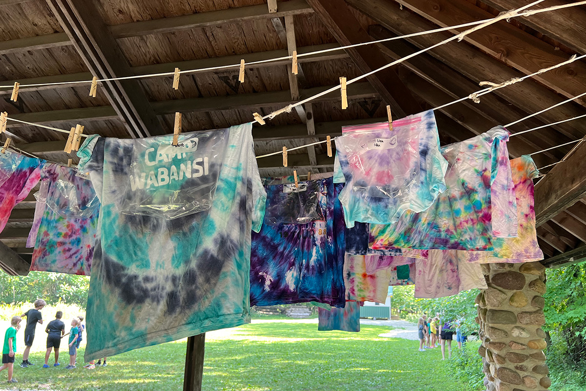 Colorful tie-dye shirts hang on a clothesline under a wooden pavilion, drying in a camp setting with children in the background.