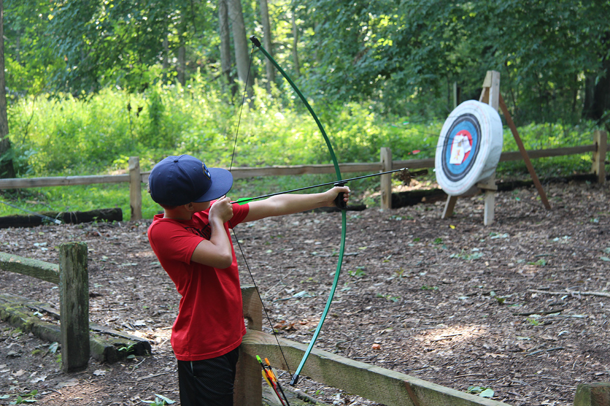 A young boy in a red shirt practices archery, aiming a bow at a target in a wooded outdoor setting.