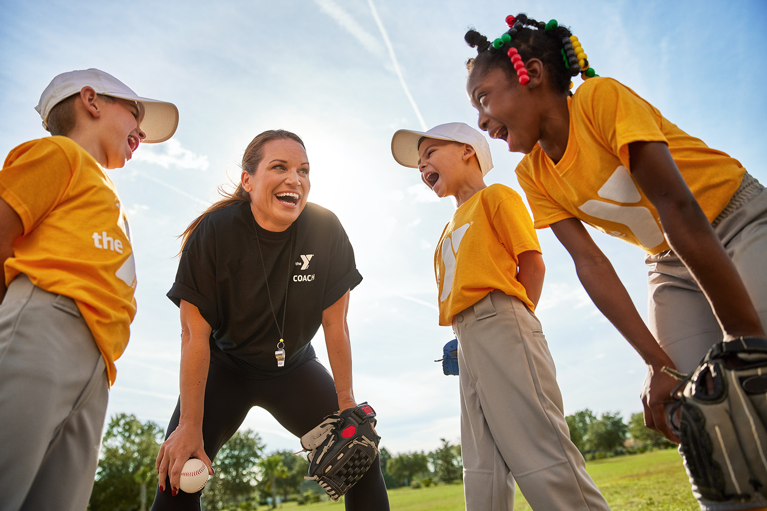 A coach smiles and talks with three children in baseball uniforms during practice on a sunny day outdoors.