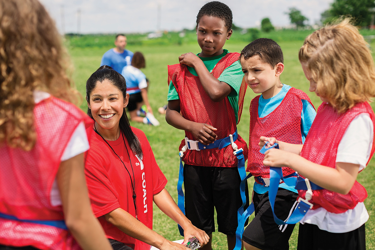 A youth sports coach talks with a group of children wearing red pinnies during a flag football game outdoors.