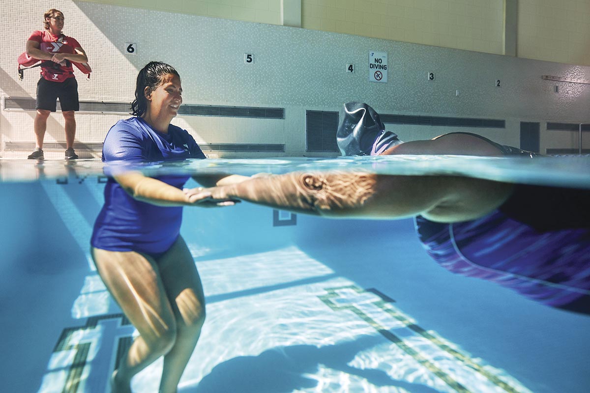 Underwater view of a swim instructor assisting a participant in a pool, with a lifeguard observing in the background.